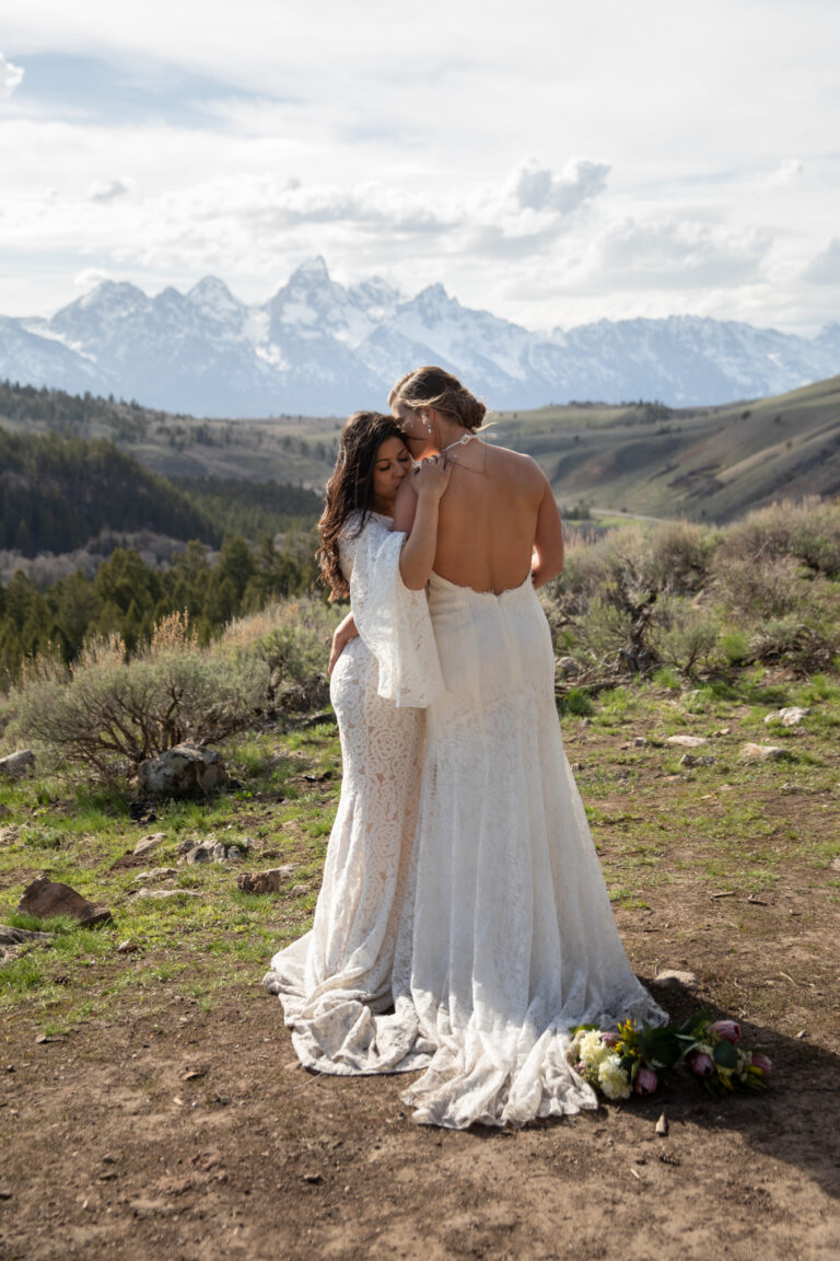 Two brides stand embracing as one gently kisses the other's shoulder.