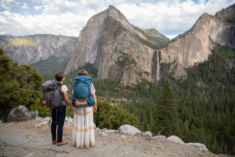 Two brides stand holding hands facing a waterfall and wearing backpacking bags and "just Married" signs hanging from the back.