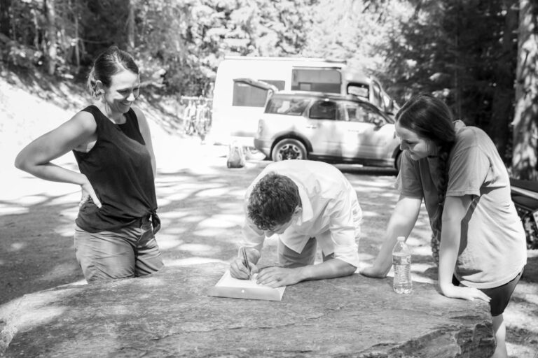 Two women watch a man signing a marriage license, using a big boulder as a table.