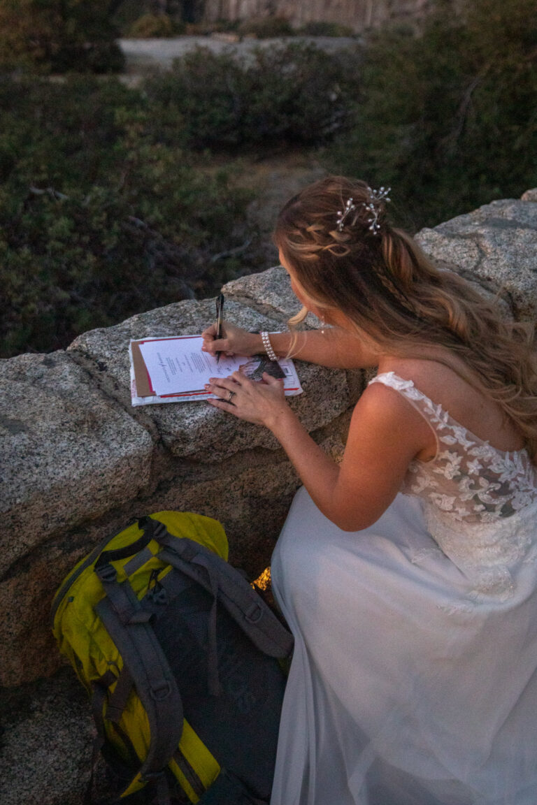 A woman kneels down using a rock wall as a table and signs her marriage license.