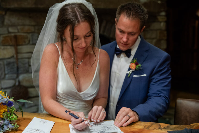 A bride holds a pen, reading a marriage license as her groom stands next to her watching.