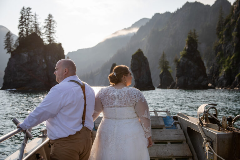 A bride and groom stand at the front of a boat in Alaska looking at the views around them.