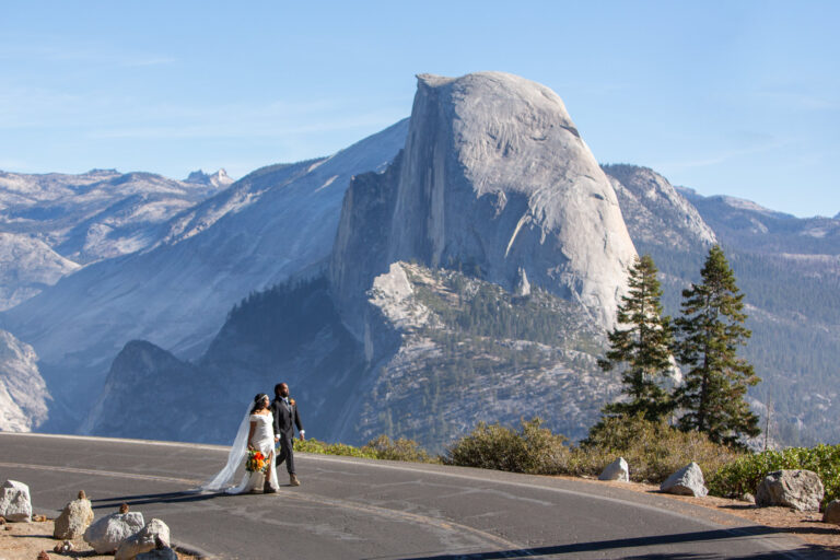 A bride and groom walk in the middle of the road in Yosemite with Half Dome behind them.