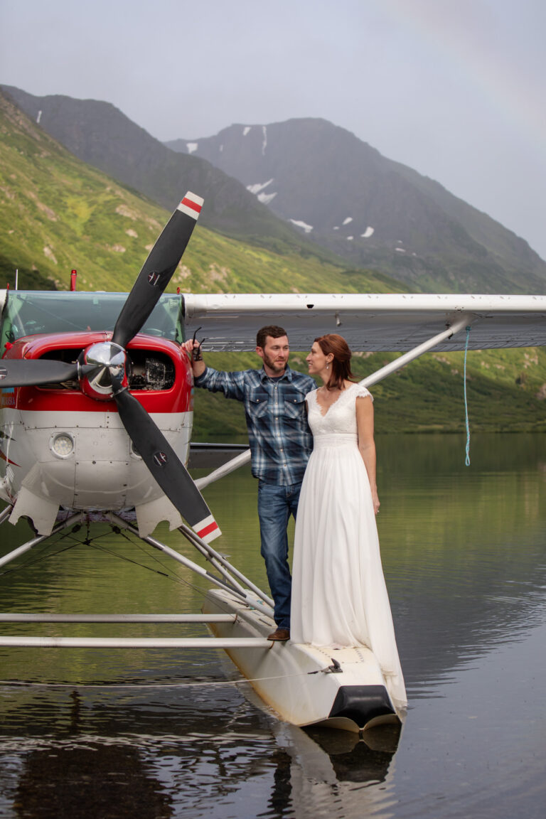 A bride and groom stand side-by-side on a floatplane that is parked in a lake in Alaska.