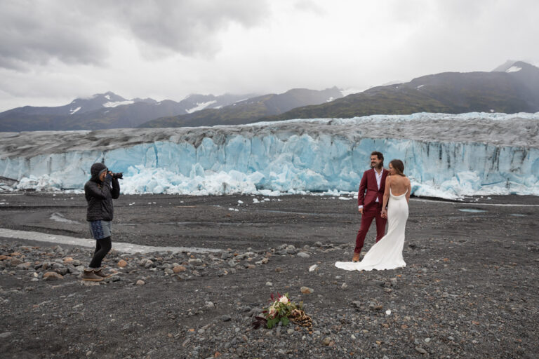 A photographer takes a photo of a bride and groom standing in front of a glacier on their elopement day.