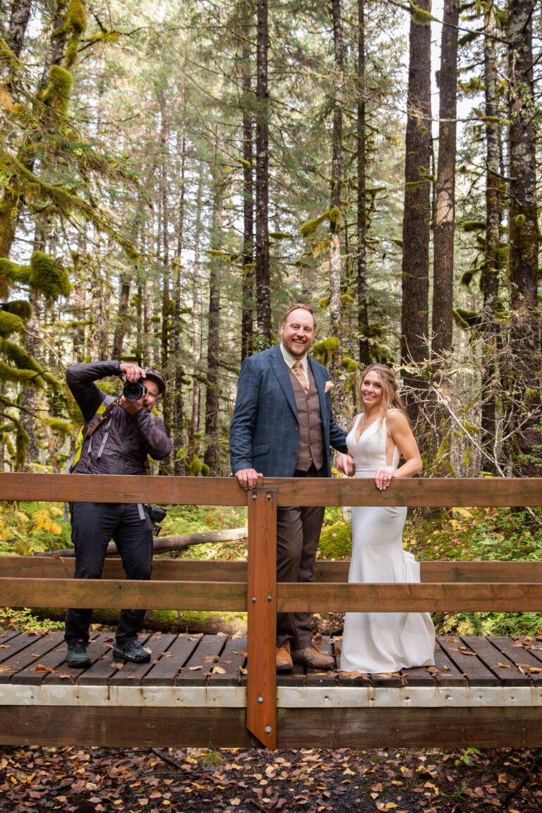 A bride and groom stand on a wooden bridge smiling as their photographer stands next to them taking a picutre.