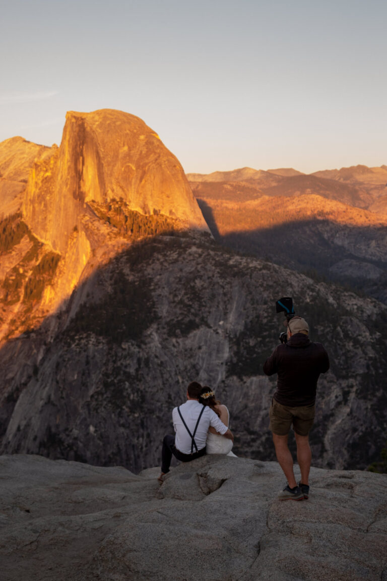 A photographer takes a picture of a bride and groom sitting on a rock in Yosemite watching the sunset on Half Dome.
