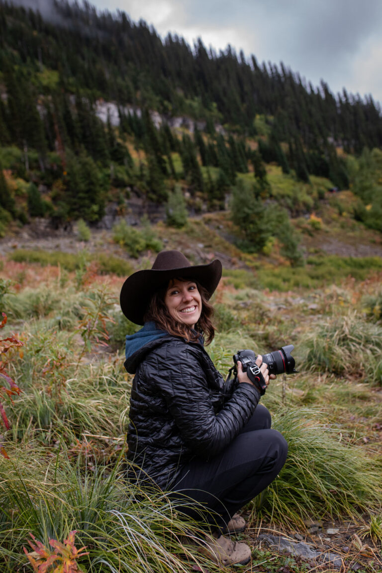 A photographer kneels down in a grassy meadow smiling and wearing a cowboy hat and holding her camera.
