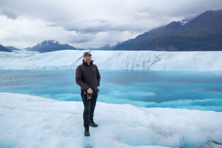 A photographer stands in front of a blue glacier pool in Alaska holding his camera and smiling.