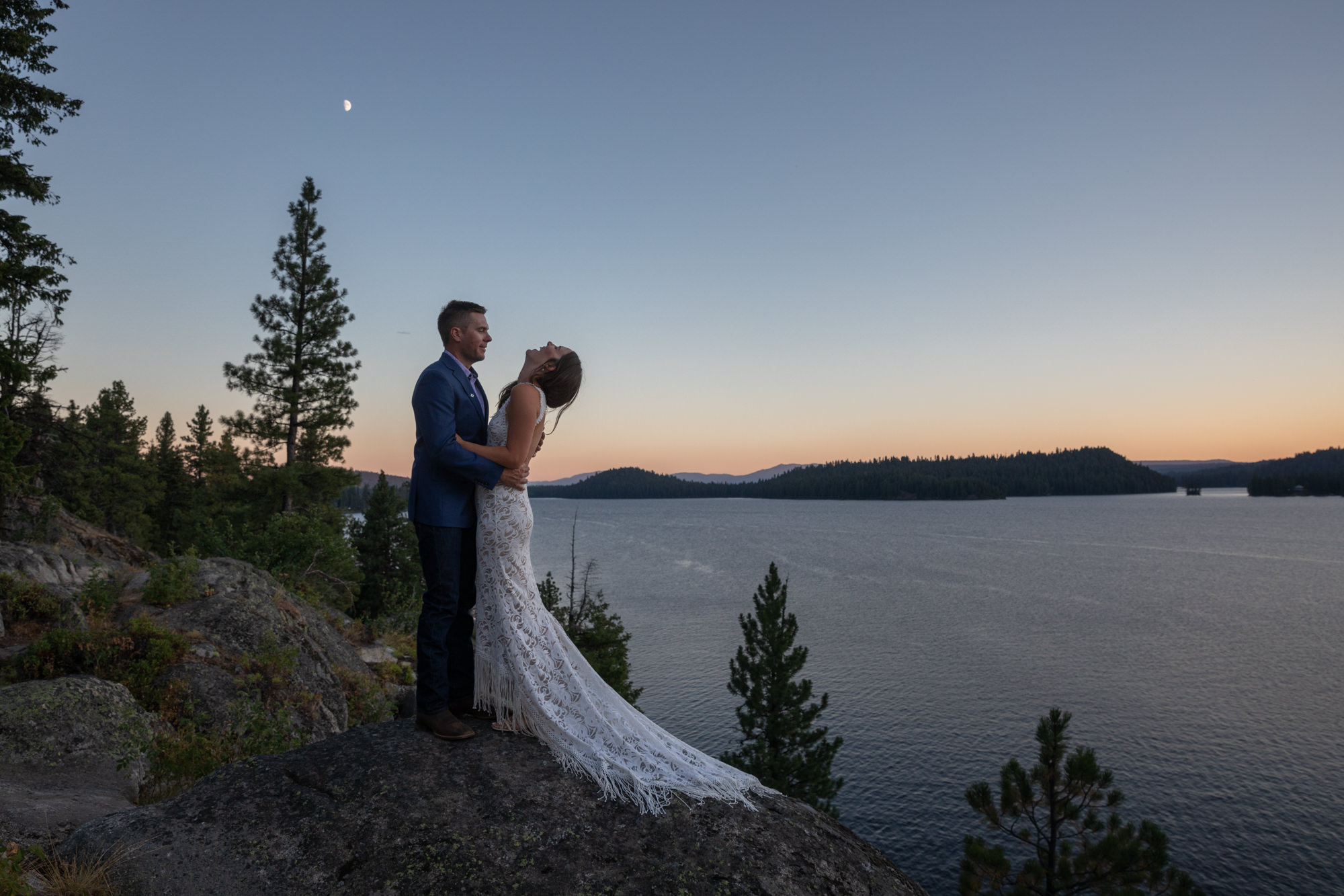 A bride throws her head back laughing as her groom holds her and they stand next to a lake after figuring out how to elope in idaho.