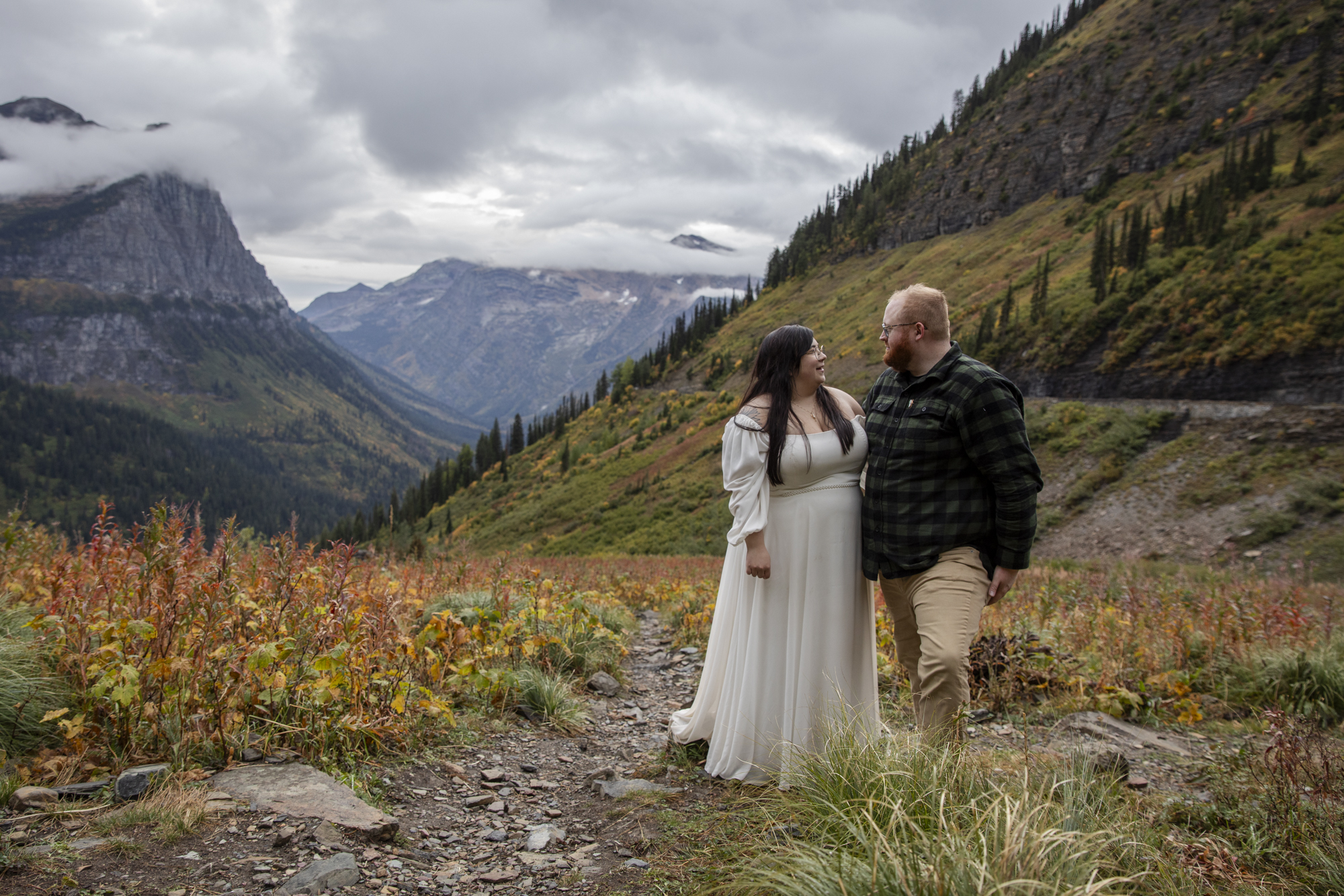 Couple walking through a mountain pass in Glacier National Park during fall, surrounded by clouds and vibrant foliage, showing what it can look like when planning how to elope in Montana.