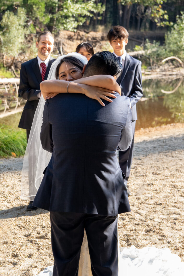 A bride hugs her groom on their intimate wedding day as her family stands behind her smiling.