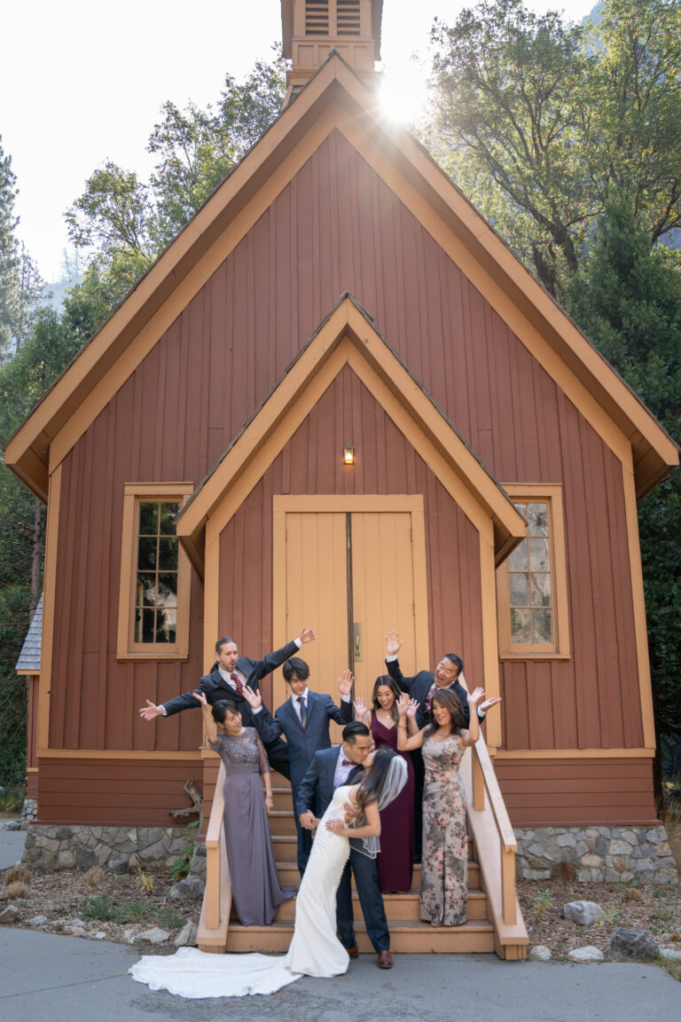 A bride and groom kiss in front of a church in Yosemite while their families stand on the stairs of the church cheering