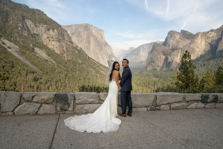 A bride and groom look back at the camera while standing at Tunnel View in Yosemite.