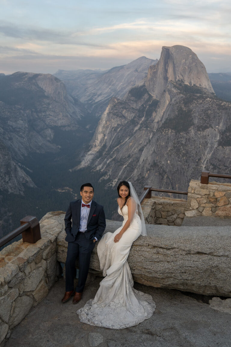 A bride and groom sit on a rock wall looking up at the camera.