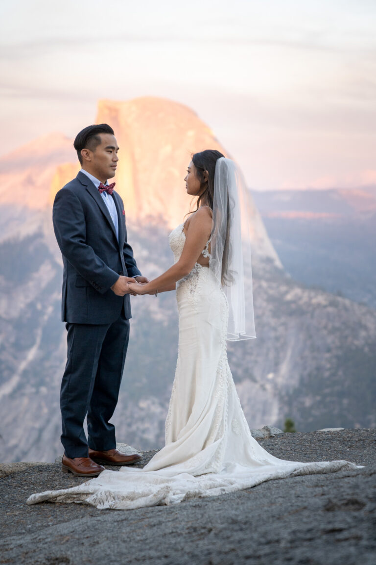 A bride and groom stand facing each other and holding hands with half dome behind them in Yosemite.