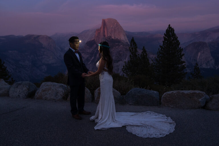 A bride and groom stand facing each other, wearing headlamps as the sky turns purple and pink around them.