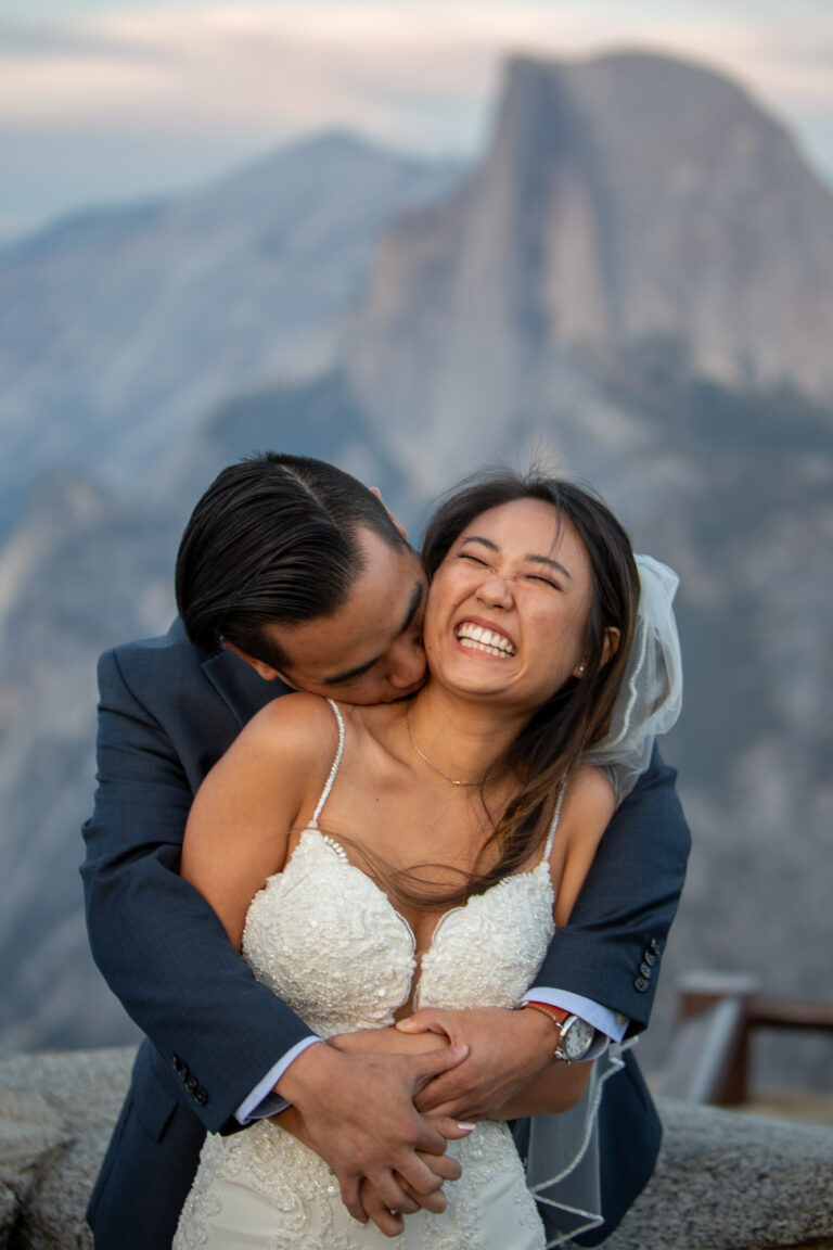 A groom stands behind is bride kissing her on the neck as she laughs and smiles.