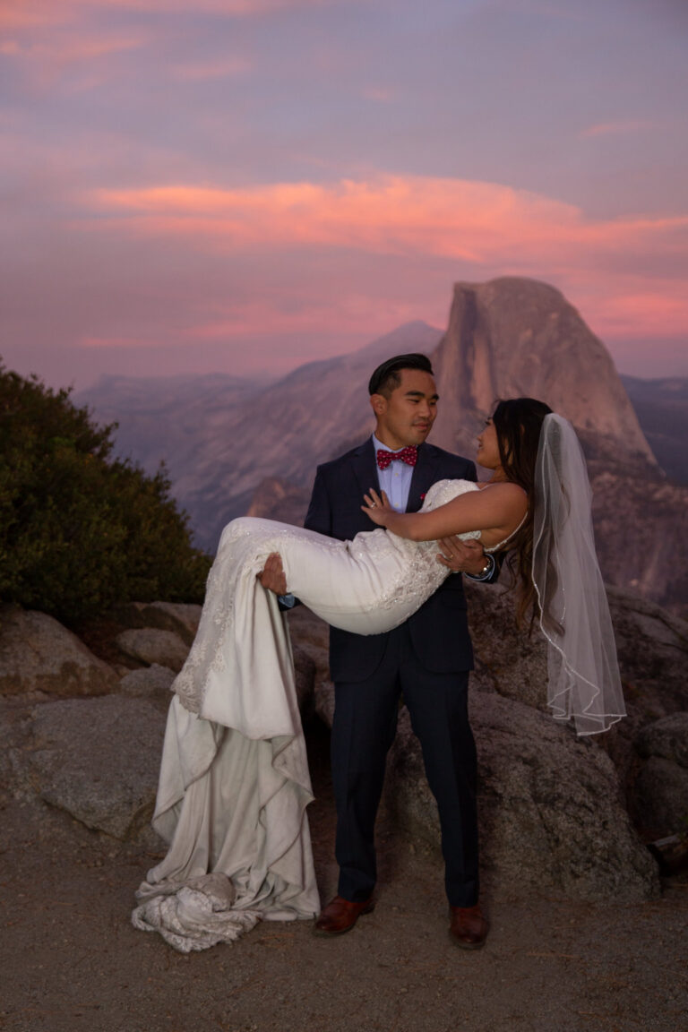 A groom holds his bride in his arms as the sun turns pink behind them.