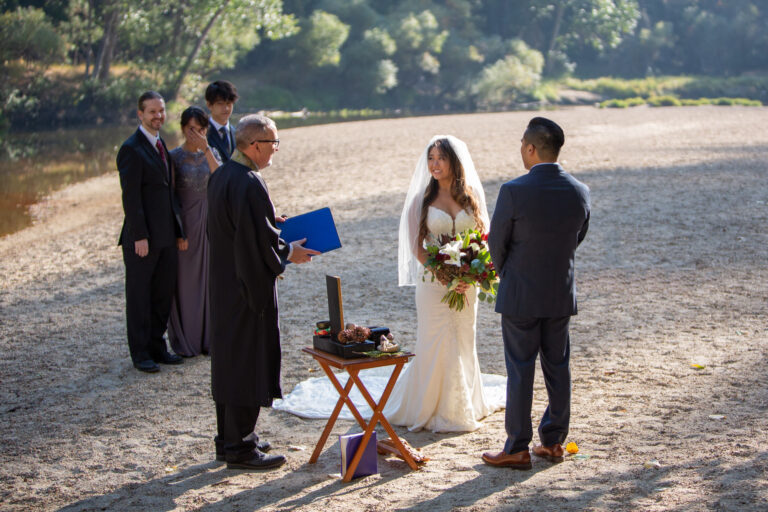 A bride and groom stand in front of an officiant on their intimate wedding day while their family looks on.