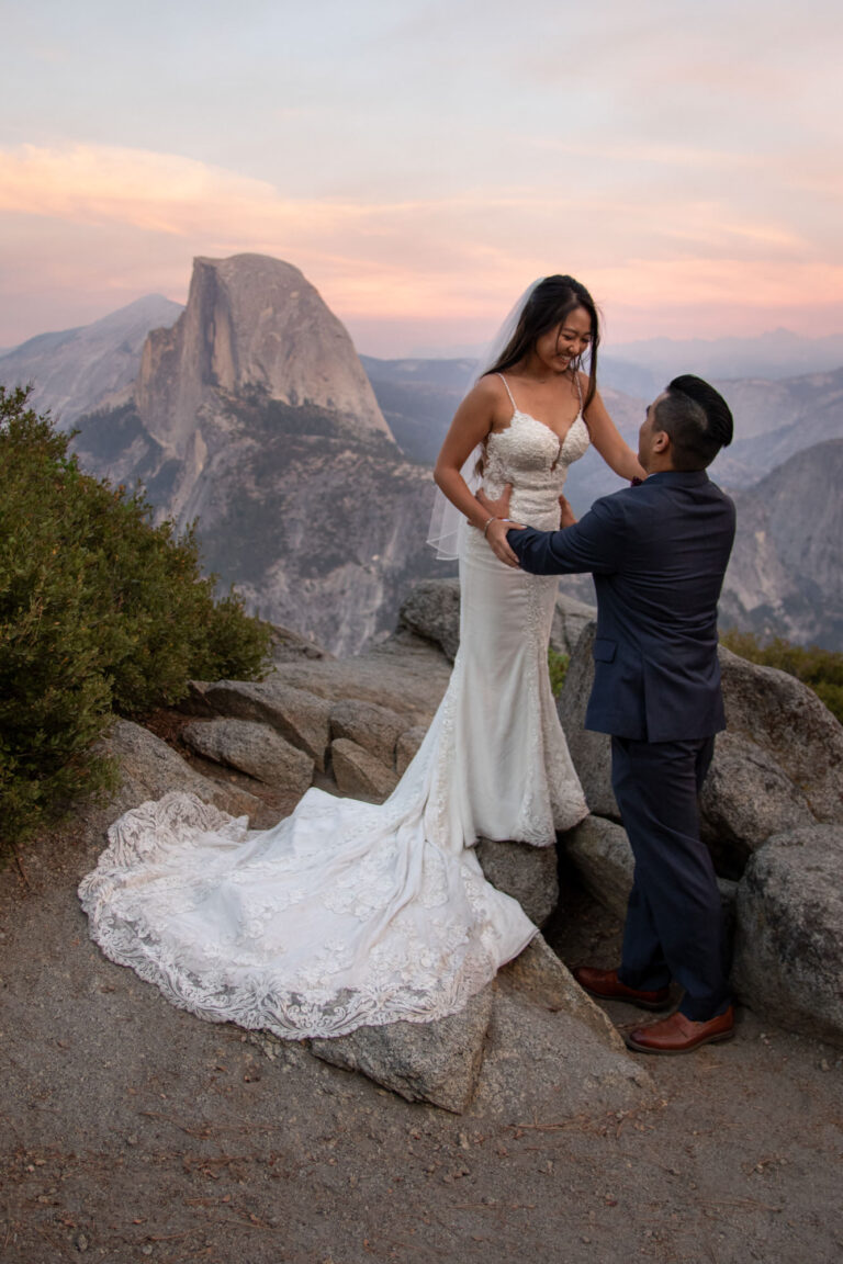 A bride stands up on a rock while her groom holds her around the waist and the sunsets behind them.