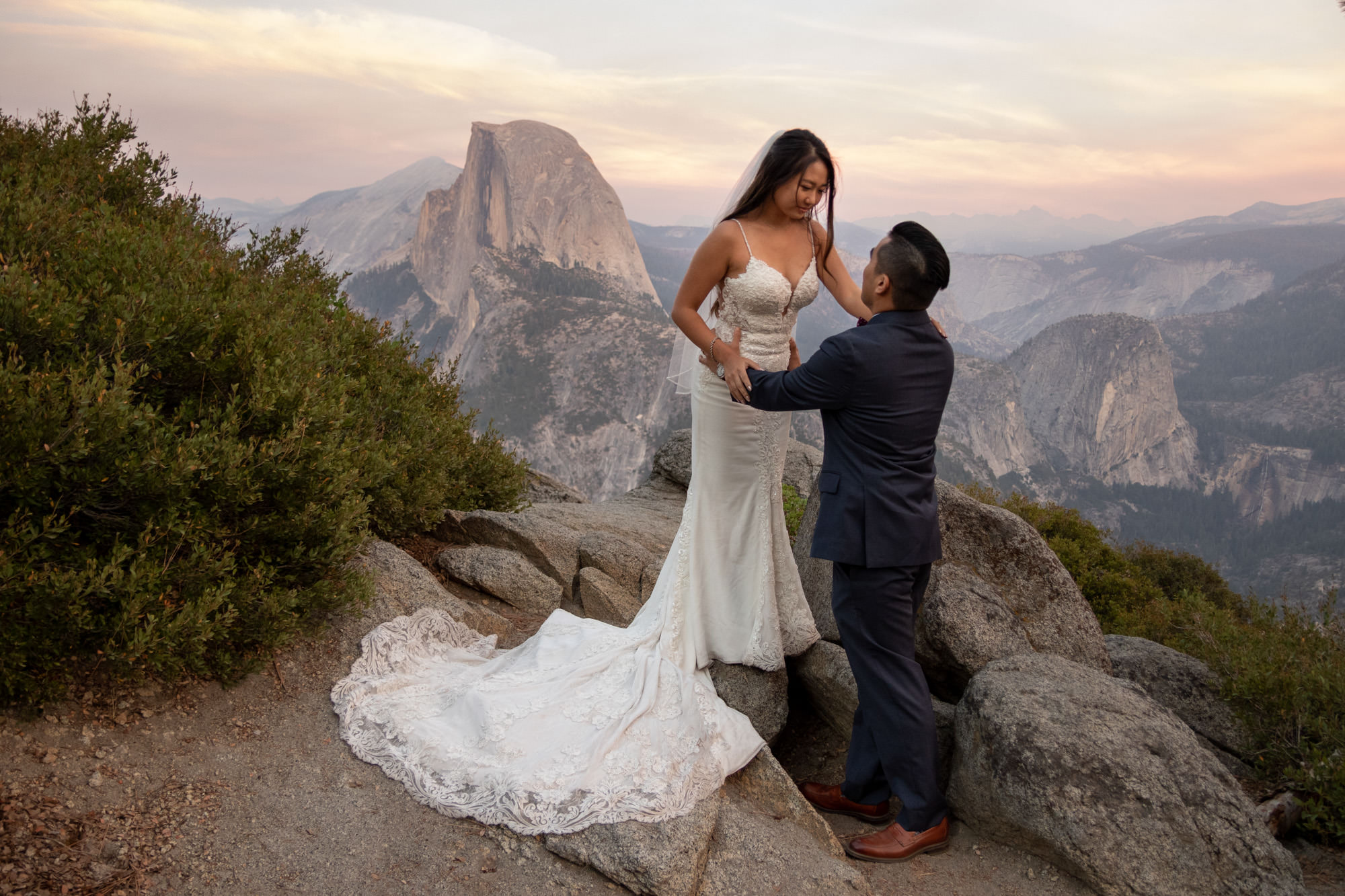 A bride stands up on a rock looking down at her groom and holding onto his shoulders on their intimate wedding day in Yosemite.