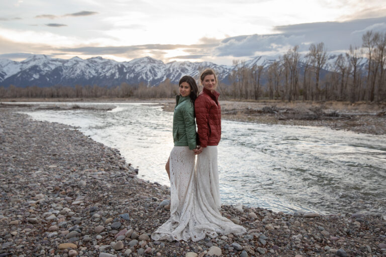 Brides stand back to back next to a river, smiling at the camera as the sunsets behind them over the mountains in Jackson Hole Wyoming