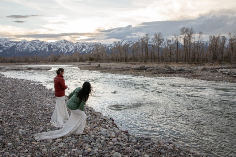Two brides stand facing a river as one bends down and throws a rock into the river.