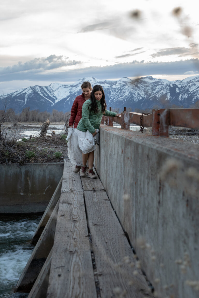 Two brides walk across a wooden bridge towards the camera as the sunsets behind them.