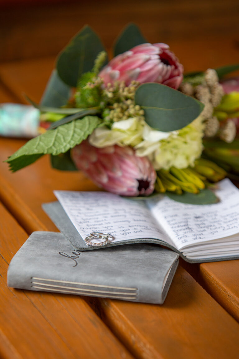 A pair of rings lie across the pages of a leather bound vow book with a bouquet of wedding flowers rests across the table.