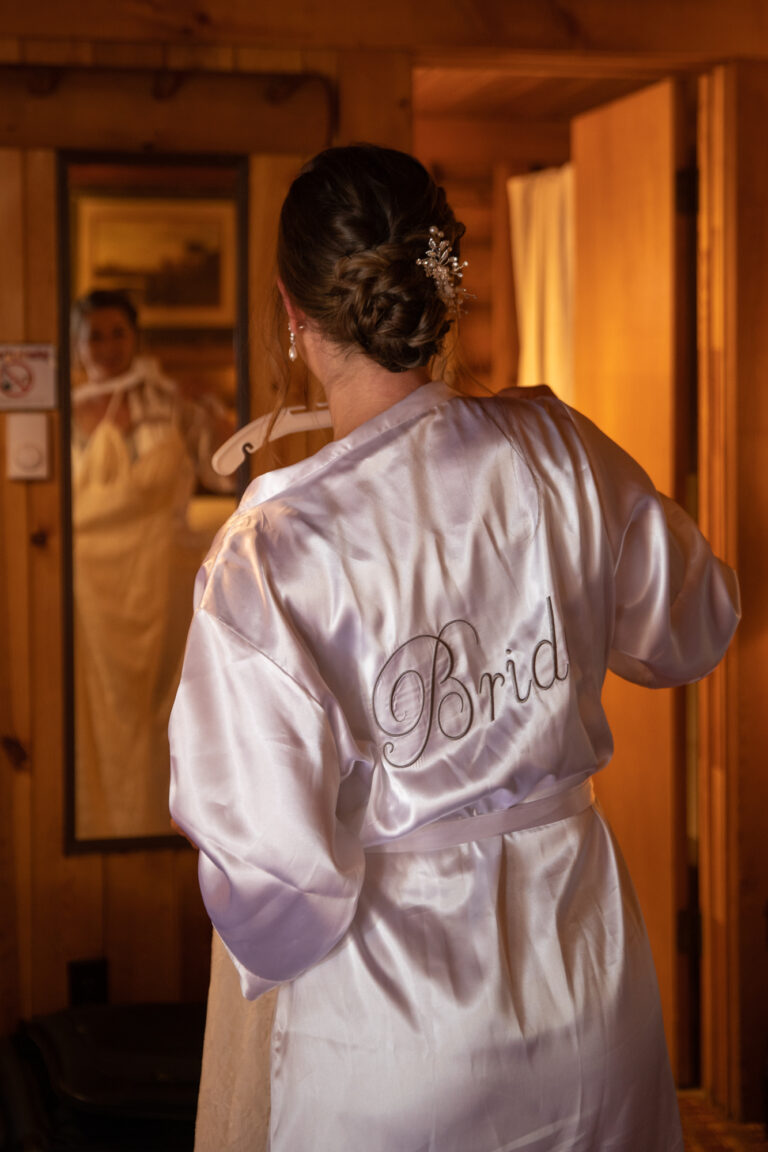 Bride stands in front of a mirror holding up her wedding dress in front of her. She's wearing a silk robe that says bride on the back.