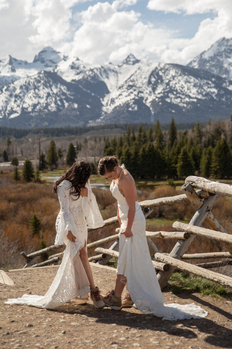 Brides stand facing each other, each holding out their foot to reveal matching hiking boots under their wedding dresses.