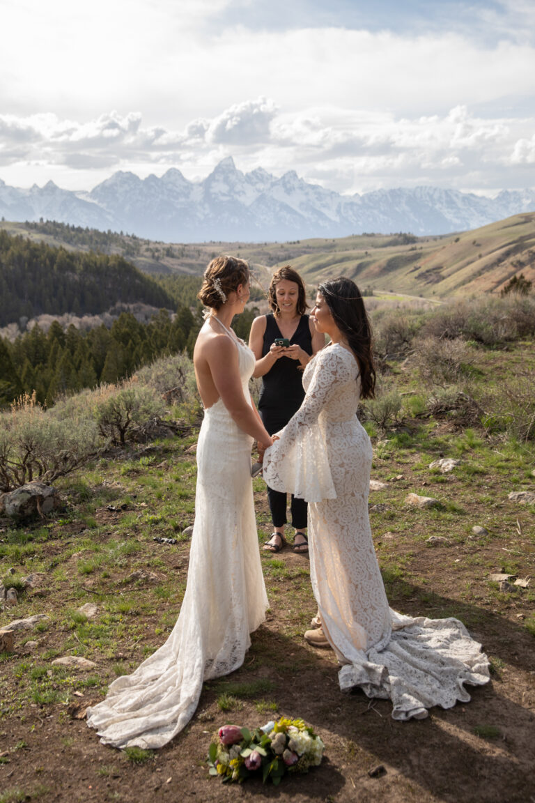 Brides stand facing each other and holding hands as the officiant stands behind them reading their ceremony script.