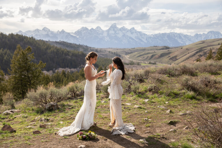 Two brides stand together on top of a mountain reading their vows to each other in Jackson Hole Wyoming.