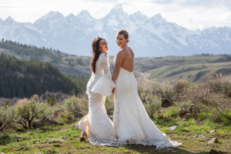 Two women stand holding hands and looking back towards the camera smiling on their wedding day with the Grand Teton mountains in front of them.