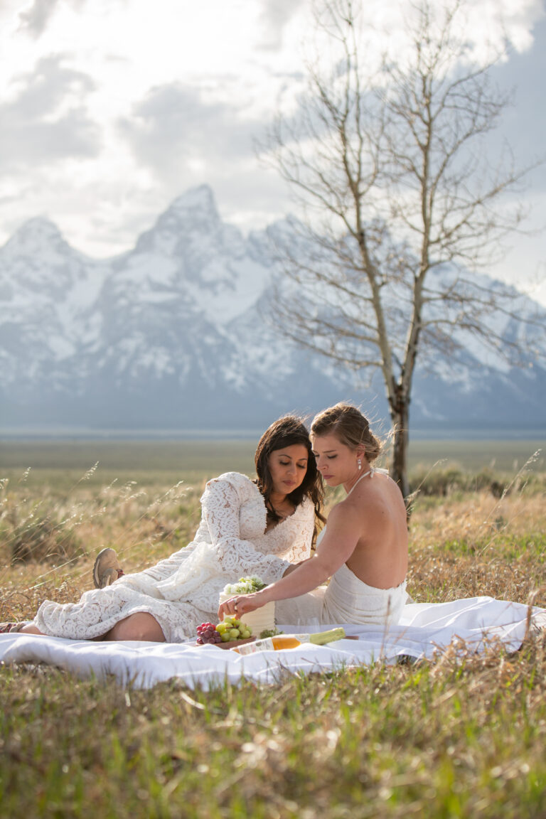 Two brides sit on a white picnic blanket eating snacks on their wedding day.