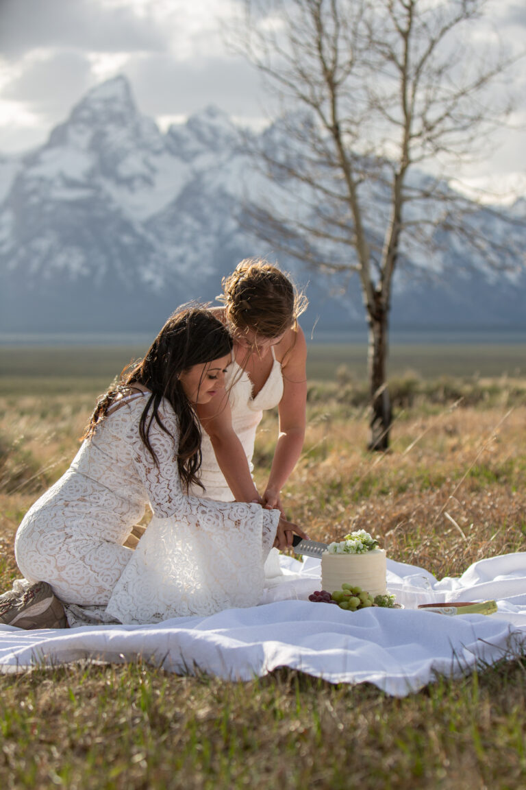 Two brides kneel on a white blanket, both holding a knife to cut their wedding cake.