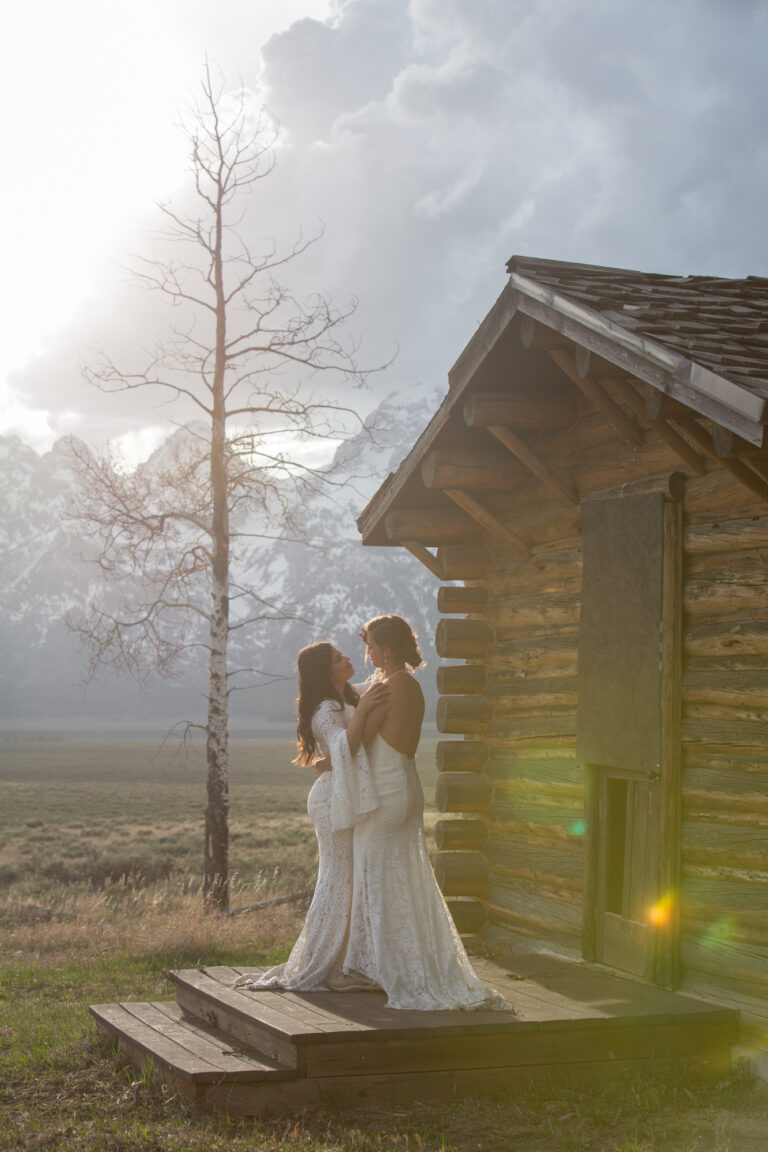 Two brides stand holding each other on the porch of a historic cabin and there is a sun flare in the bottom right corner of the photo.