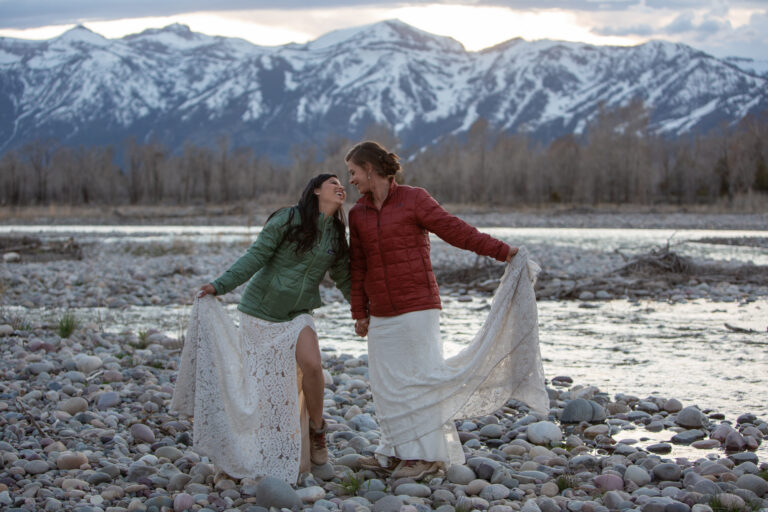 Two brides stand kissing in the middle and holding their wedding dresses out to the side next to a river in Wyoming after eloping in Grand Teton