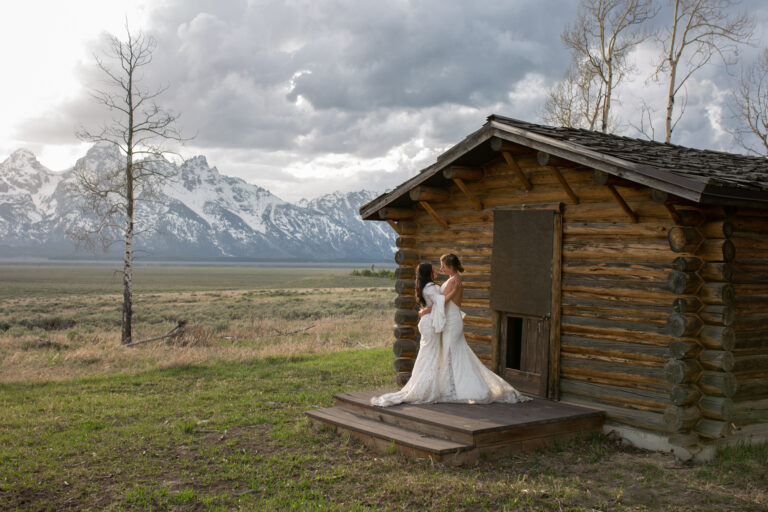 Two brides stand on the porch of a historic cabin in Grand Teton National Park, sharing their first dance as a married couple.