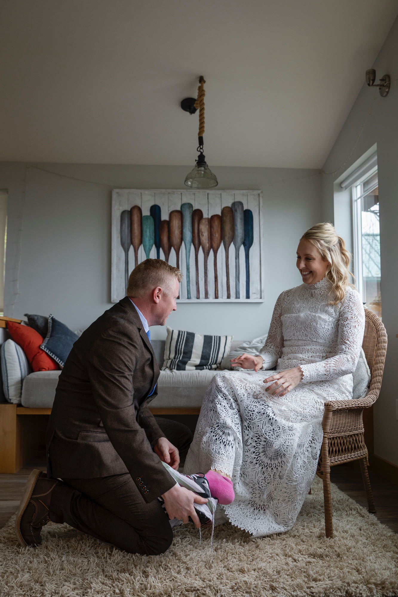 A groom kneels down on the floor, helping his bride put on her shoes as she sits in a chair.