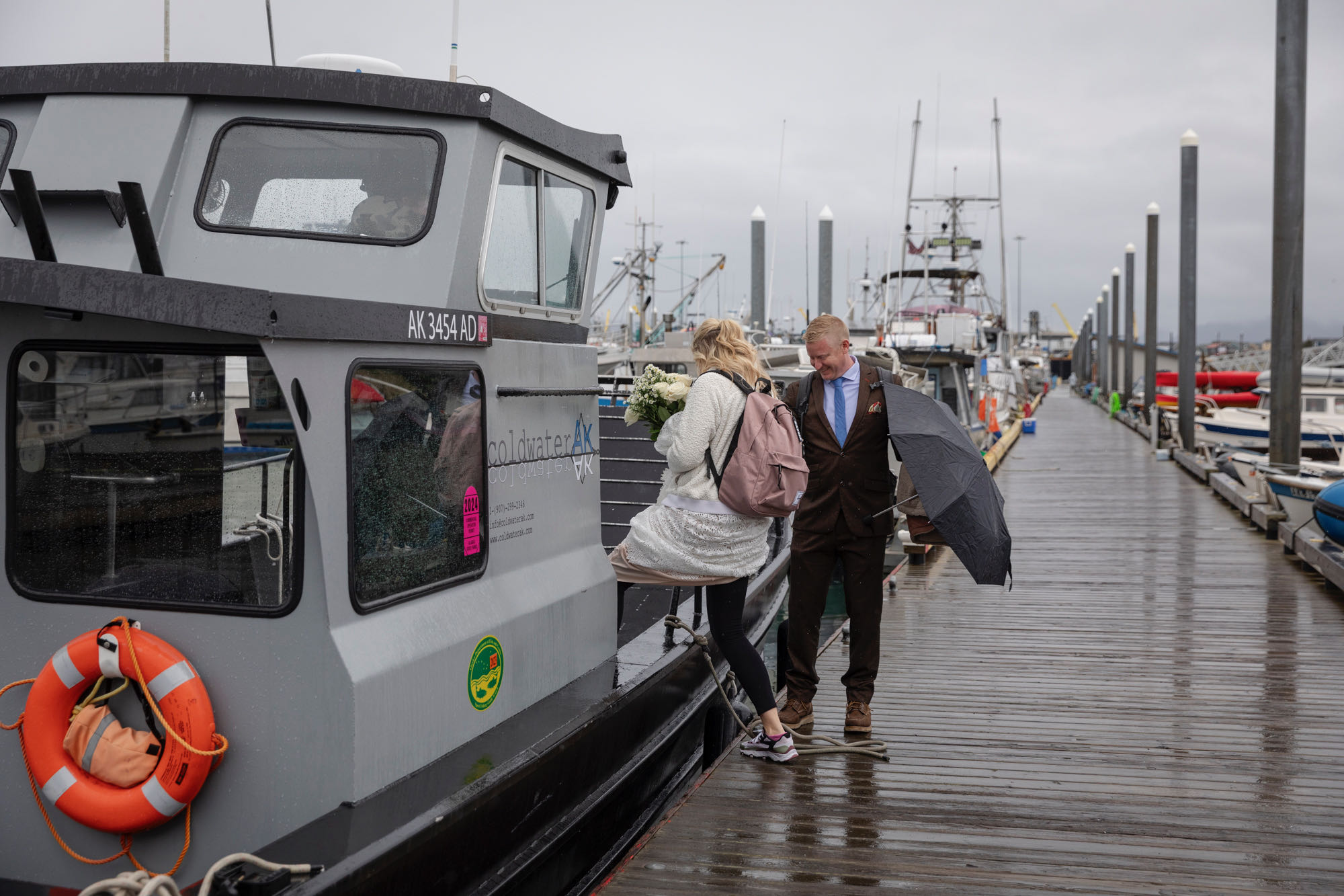 A bride sets onto a boat in Kachemak Bay, Alaska on her wedding day, while her groom stands on the dock, helping her.