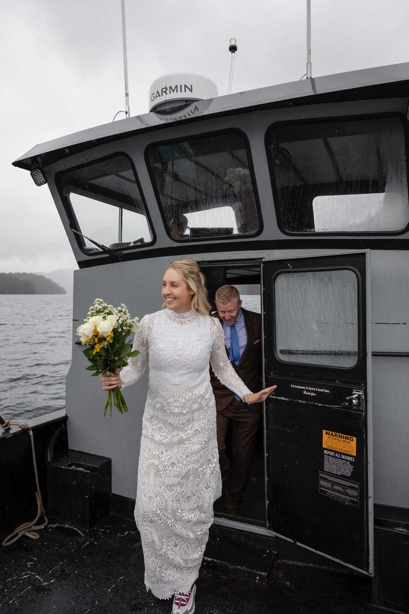 A bride wearing a white lace dress and holding a bouquet of flowers walks out a door on a boat smiling.