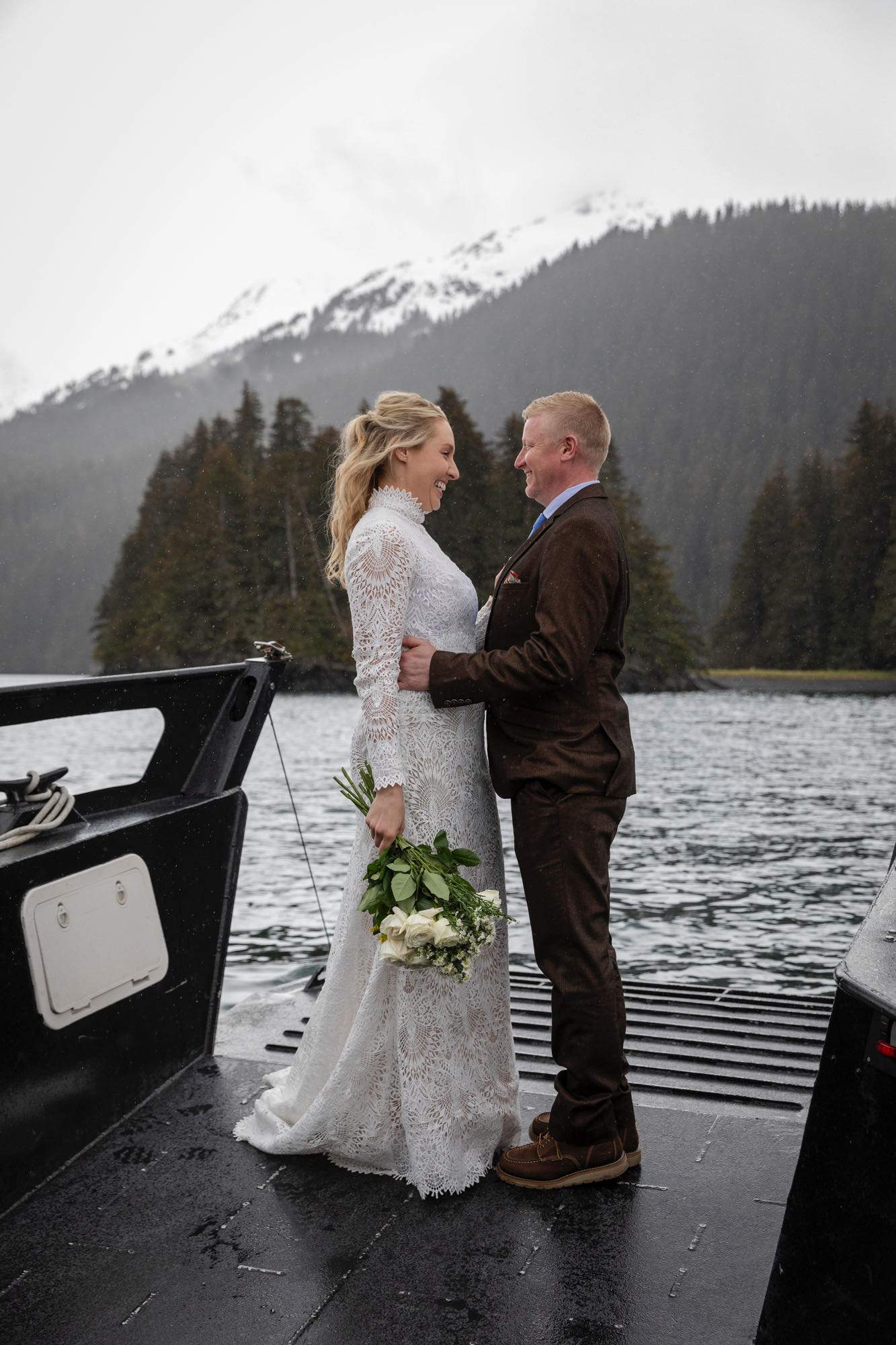 A bride and groom stand facing each other smiling on the bow of a boat. 