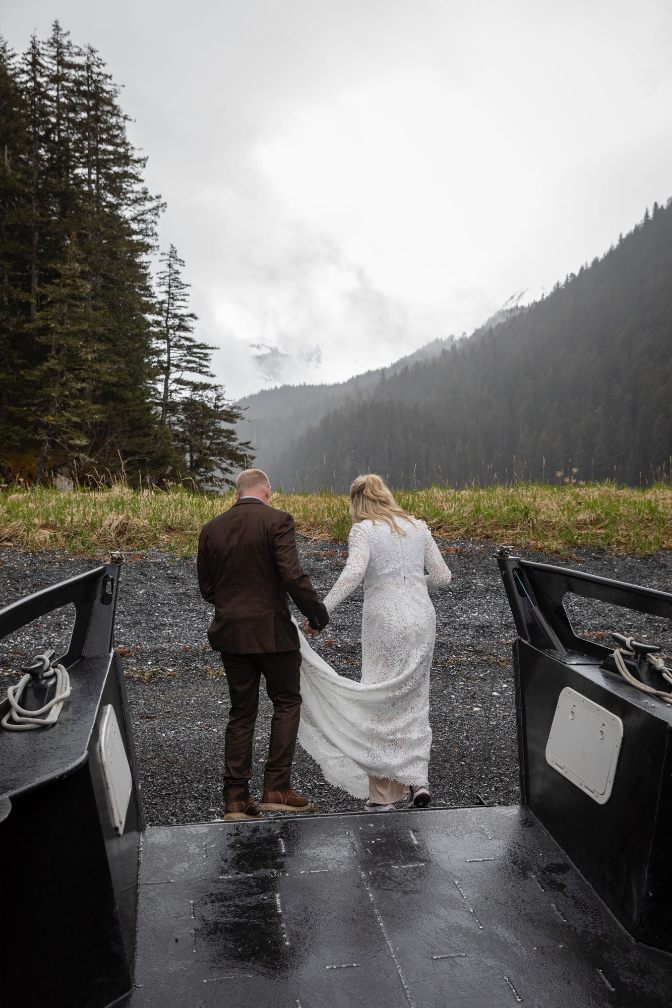 A bride and groom walk off the front of a docked boat onto a rocky black sand beach in Kachemak bay.