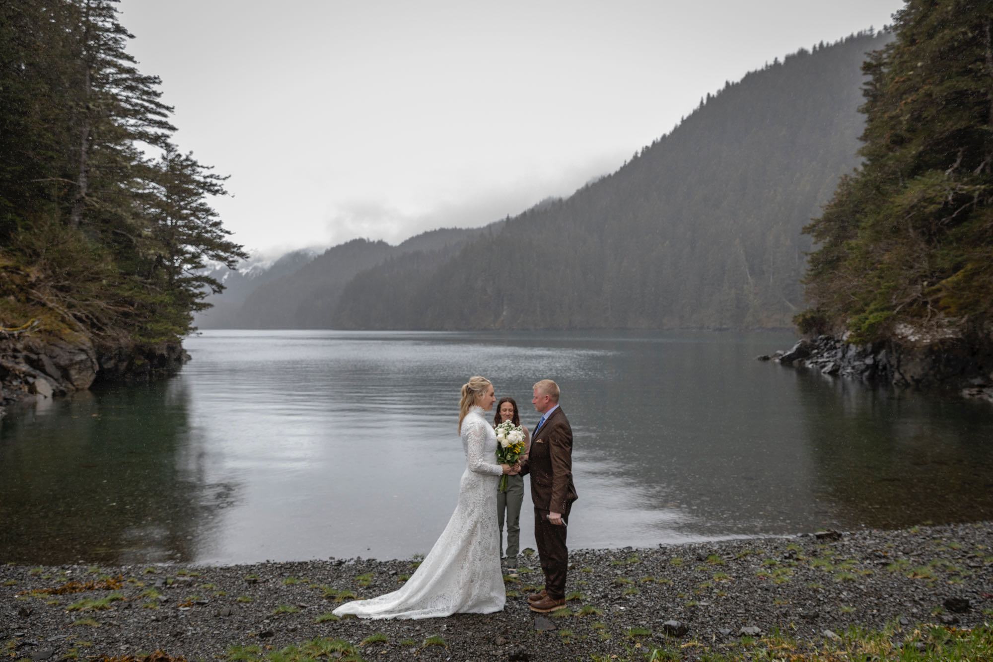 Couple exchanging vows on rocky shoreline during Kachemak Bay elopement with misty mountains in background