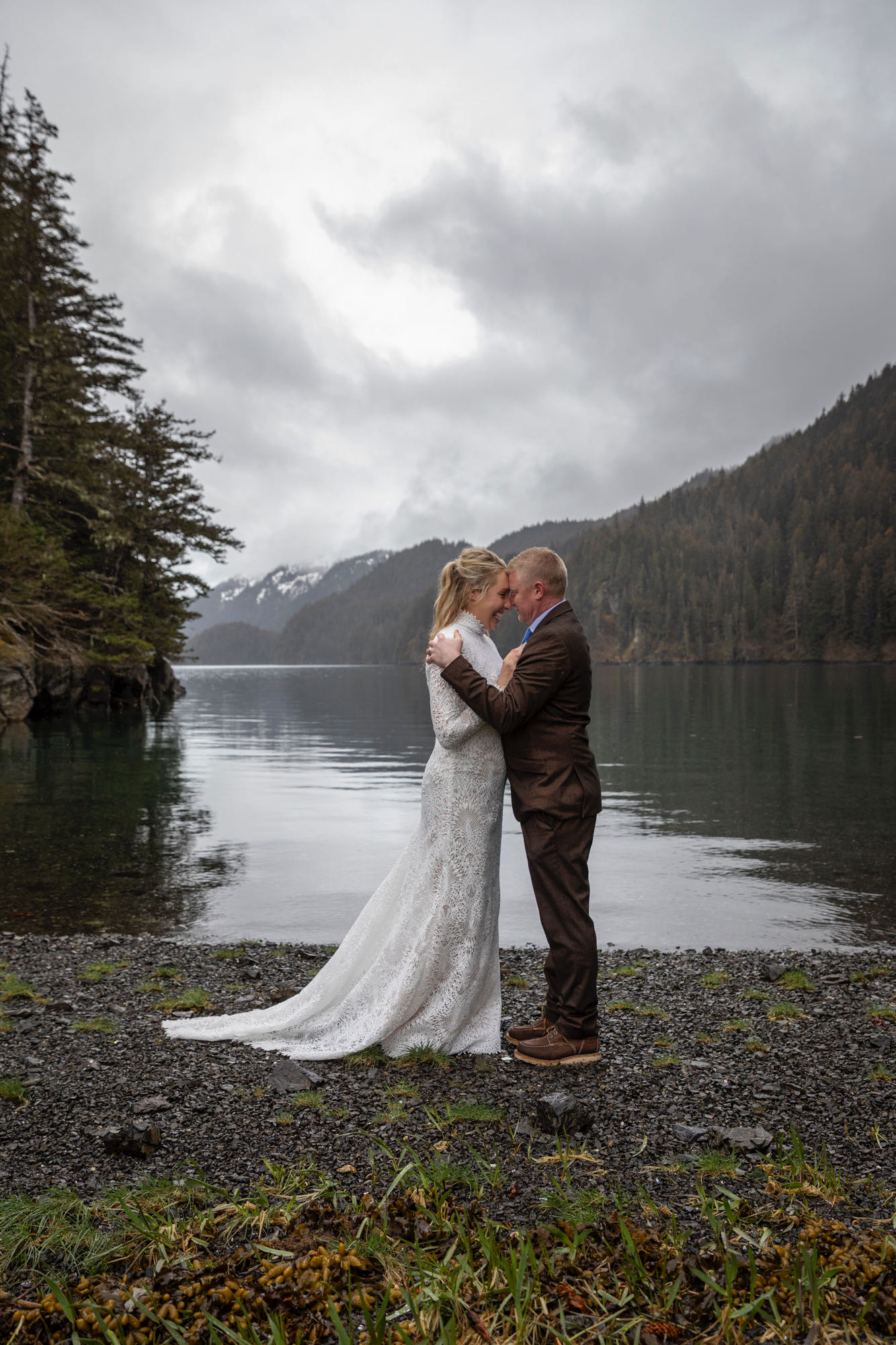 Bride and groom embracing on quiet beach during rainy Alaska elopement with mountains and water behind them