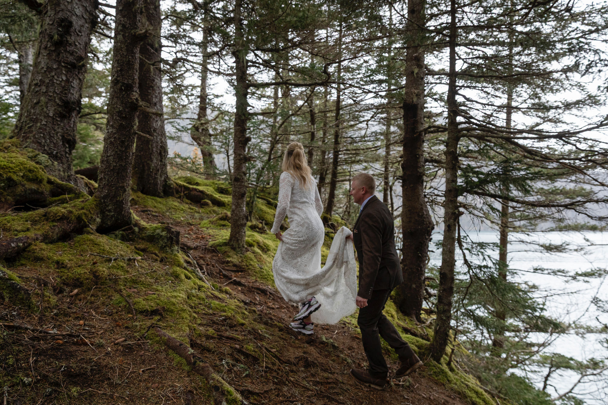 Bride and groom walking through mossy forest overlooking the water during Alaska elopement