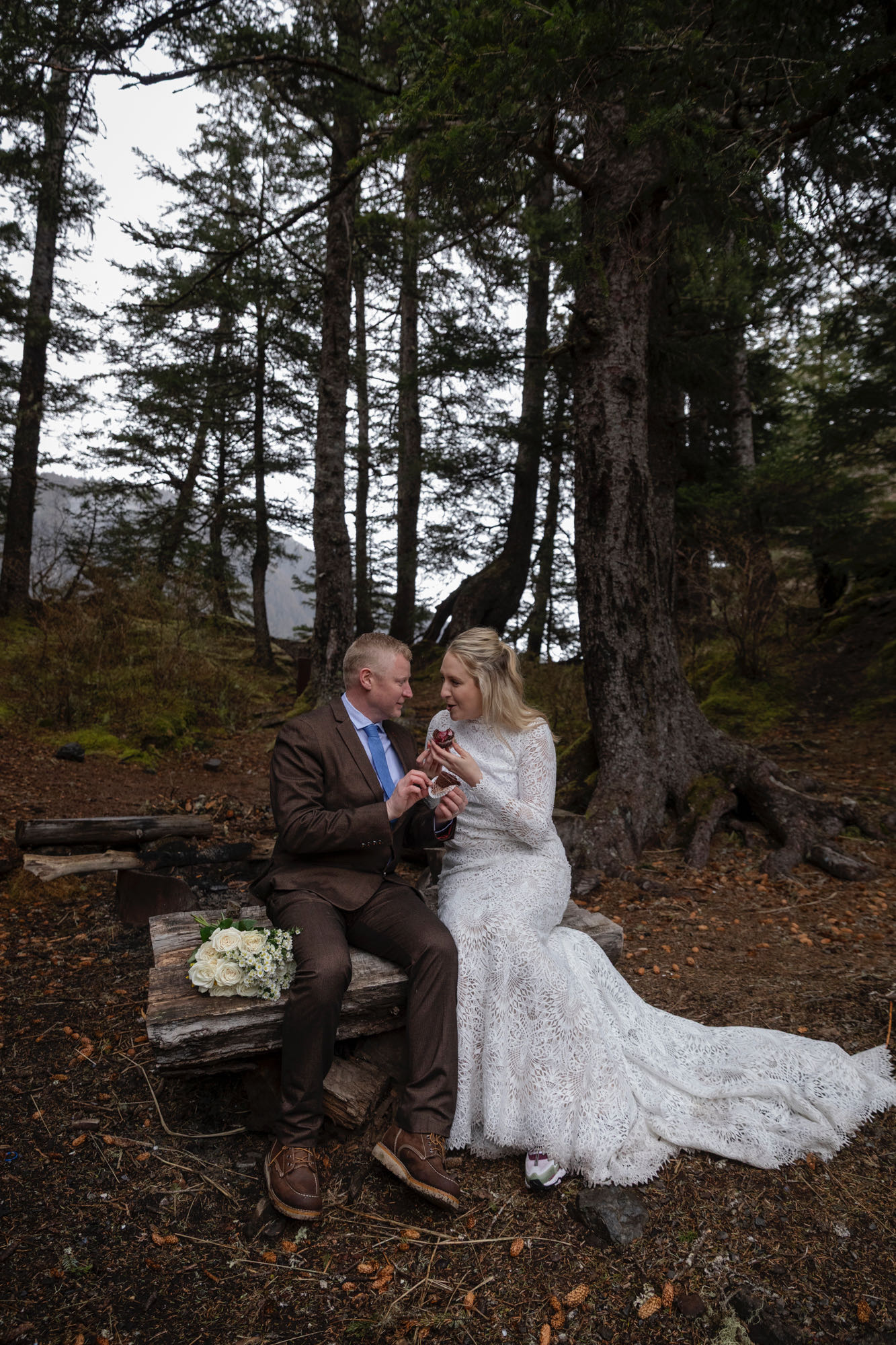 Bride and groom sitting on log in forest sharing cupcakes during relaxed Kachemak Bay elopement