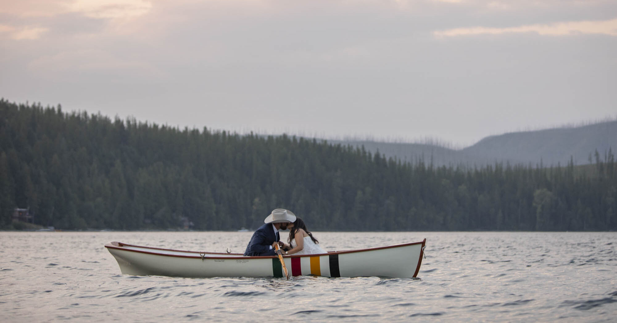 A bride and groom sit in a white canoe in the middle of a lake kissing after their Lake McDonald wedding.